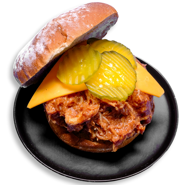 A hearty open-faced BBQ chicken sandwich rests on a black plate, featuring shredded chicken thighs drenched in smoky tomato-based barbecue sauce, layered with a thick slice of cheddar cheese and crowned with crisp, crinkle-cut pickles, all nestled on a whole grain Dave’s Killer Bread bun. This colorful dish is a bold and satisfying healthy prepared meal packed with protein and whole food ingredients.