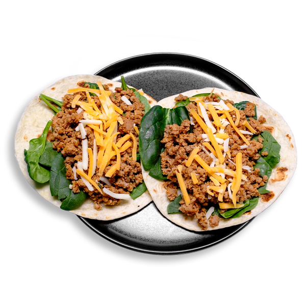 Two open-faced tacos sit on a black plate, each built on a lightly toasted flour tortilla and generously filled with vibrant green spinach, spiced lean ground pork seasoned with a custom Jamaican jerk blend, and a colorful sprinkle of Cheddar Jack cheese.