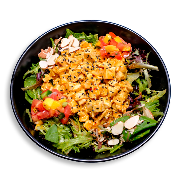 A lively and colorful healthy meal prep plate of salad featuring a generous portion of diced chicken breast tossed in a golden, creamy dressing and sprinkled with black sesame seeds. The chicken sits atop a lush bed of mixed greens, including kale and spinach, creating a fresh, earthy base. Around the edges, vibrant accents of diced mango, fire-roasted red peppers, roma tomatoes, and corn add a pop of color, while thinly sliced almonds lend a satisfying crunch.