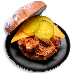 A hearty open-faced BBQ chicken sandwich rests on a black plate, featuring shredded chicken thighs drenched in smoky tomato-based barbecue sauce, layered with a thick slice of cheddar cheese and crowned with crisp, crinkle-cut pickles, all nestled on a whole grain Dave’s Killer Bread bun. This colorful dish is a bold and satisfying healthy prepared meal packed with protein and whole food ingredients.