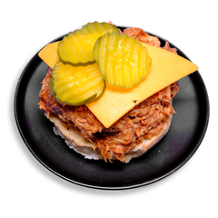 A visually striking, protein-packed healthy prepared meal featuring tender chicken thighs slow-cooked in tangy barbecue sauce, stacked over a whole grain burger bun. The cheddar cheese slice gently melts into the savory meat, while 3 thick-sliced pickles add a sharp crunch, making this sandwich a satisfying fusion of flavor, texture, and nutrient-dense ingredients.