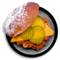 A healthy prepared meal served on a sleek black plate: pulled barbecue chicken thighs sit atop a hearty Dave’s Killer Bread bun, layered with a sharp slice of cheddar cheese and crowned with 3 crunchy pickles. This visually inviting sandwich offers bold flavor and clean ingredients, ideal for anyone looking for health food that doesn’t sacrifice taste.