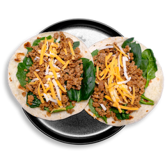 A pair of health-forward jerk pork tacos are presented on a dark plate—each tortilla layered with tender spinach leaves, all-natural ground pork infused with savory Creole and Jamaican jerk seasonings, and topped with shreds of golden cheddar and creamy white Jack cheese. 