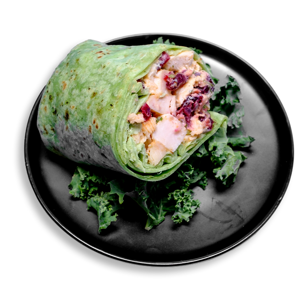 A vibrant health food wrap featuring tender diced Creole-seasoned chicken, tangy Greek yogurt, and sweet dried cranberries, all wrapped in a toasted spinach tortilla. Hints of fresh celery and aromatic tarragon peek through, with the wrap cut in half and nestled on a bed of leafy kale—an ideal healthy meal prep for those seeking flavor-packed nutrition.