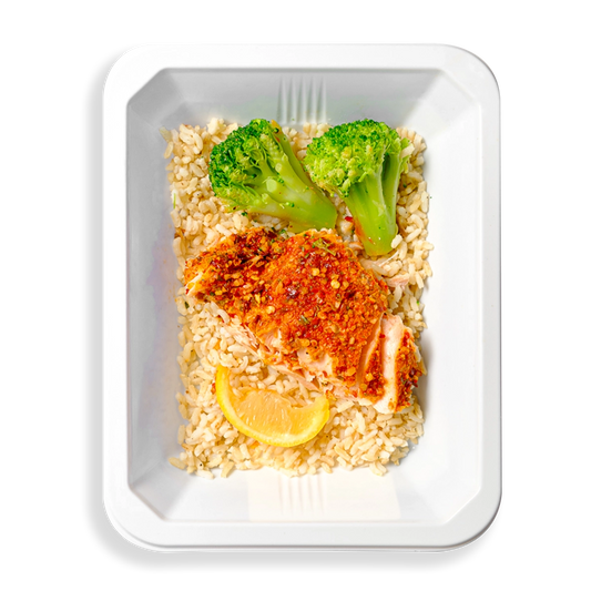 The salmon sitting on top of a bed of brown rice looks extremely flavorful based on the seasonings it has on its surface.  Accompanying the salmon in this white container are two large, green pieces of broccoli and a lemon wedge.  This meal looks like it is designed to be both extremely flavorful and healthy.  This prepared meal is in a traveling container making me think this is for health conscious individuals having healthy prepared food delivered to their homes.