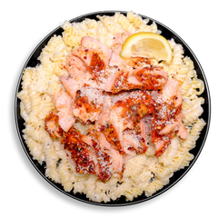 A black plate heaped with rotini-style brown rice pasta, soaked in a velvety Alfredo sauce made from heavy cream and parmesan. Atop the pasta lies a colorful spread of shredded Atlantic salmon, richly seasoned with lemon, roasted garlic, and Creole spices. Finished with a wedge of lemon, this healthy prepared meal delivers both comfort and clean nutrition.