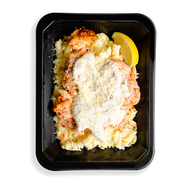 A pale, creamy salmon pasta with flakes of spice-rubbed Atlantic salmon over brown rice noodles, topped with parmesan and a light Alfredo sauce filling the black togo container.  This looks to be a hearty prepared meal designed for health conscious people who love eating.