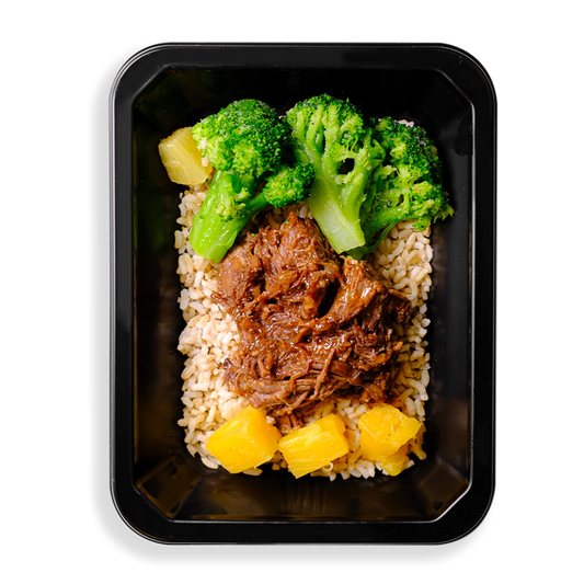 Vibrant green broccoli pulled beef and pineapple chunks sit on top of a bed of brown rice in a black to-go food container.  This meal is obviously focused on being extremely healthy while not sparring flavor.  The container that this food is in must be for a health food delivery services or a store where you can pick up healthy prepared meals.