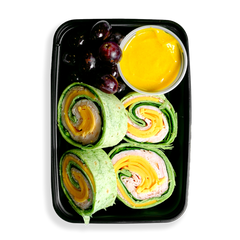 A turkey roll up cut into four even pieces alongside a yellow sauce and red grapes fill this container.  This looks like a classic healthy lunch designed to be both flavorful and healthy.  The to-go container that this food is photographed in looks like it is either ready for delivery or instore pickup but is clearly food for people eating on the go but do not want to sacrifice good health for convenience. 