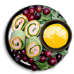 Healthy meal prep platter featuring colorful spinach wrap pinwheels stuffed with layers of smoked turkey, cheddar slices, and crisp spinach. Juicy red grapes dot the leafy green garnish, creating a bright contrast against the black plate. A smooth pool of yellow mustard dipping sauce adds tangy flavor to this protein-rich, low-nitrate health food selection that blends fresh ingredients with visual appeal.
